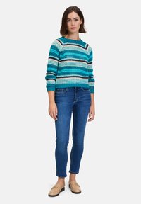 Pull en tricot avec des rayures turquoise, bleues et crème ; encolure ronde ; manches longues. Associé à un jean moulant bleu foncé et des mocassins beiges.