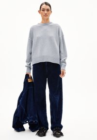 Frau trägt einen grauen Pullover und eine dunkelblaue Samthose und hält eine passende dunkelblaue Samtjake gegen einen weißen Hintergrund.