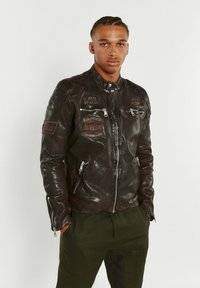 Veste de moto en cuir noir avec des accents matelassés aux épaules, des zips en métal argenté et des patches brodés sur le devant et les manches.