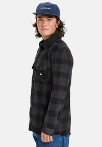 Camisa de franela a rayas en negro y gris con diseño de botones y bolsillo en el pecho. Textura suave, mangas largas, cuello redondo y acento de logo.