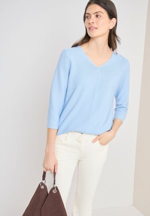 Femme en pull bleu clair à col en V et pantalon blanc tenant un sac à main marron, debout devant un fond clair uni.