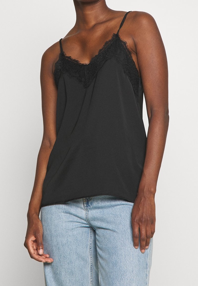 Camisole noire avec bordure en dentelle le long du décolleté, bretelles spaghetti réglables et coupe décontractée. Associée à un jean bleu clair.