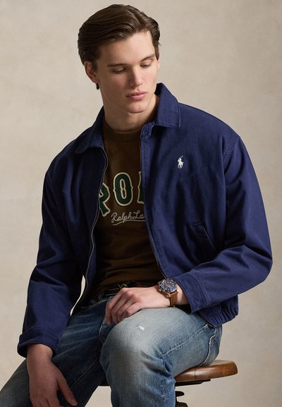 Mladi moški sedi na stolčku, oblečen v mornarsko modro jakno, rjavo Polo Ralph Lauren majico, svetlo modre jeans hlače in ročno uro, gleda dol.