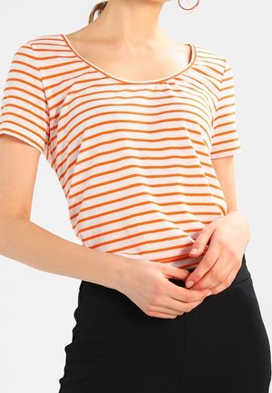 Kvinde iført en hvid kortærmet skjorte med orange vandrette striber og sorte bukser, hænderne samlet ved taljen.