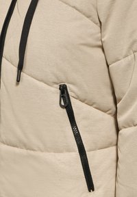 Beige polstret jakke med et diagonalt quiltet mønster, der har en sort lynlåslomme og mørke snore. Glat stofstruktur.