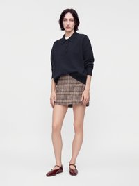 Femme portant un polo à manches longues noir, une mini-jupe beige à carreaux et des chaussures Mary Jane bordeaux, se tenant devant un fond blanc uni.