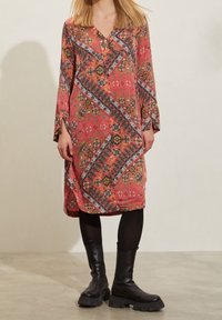 Vestido rojo con un patrón, de mangas largas, escote en V y detalles en negro. Presenta diseños florales y geométricos, combinado con botas negras hasta el tobillo.
