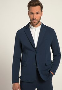 Navyblazer mit tailliertem Schnitt, Ein-Knopf-Verschluss, Reverskragen, zwei vorderen Taschen und glatter Textur, kombiniert mit einem weißen Hemd.