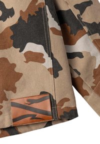 Il tessuto in cotone con motivo mimetico presenta colori beige, marrone e nero. È visibile un'etichetta in pelle marrone con dettagli di design neri.