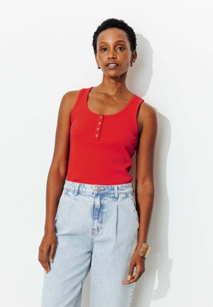 Femme aux cheveux courts et bouclés portant un haut rouge sans manches avec des boutons et un jean taille haute bleu clair, debout devant un fond blanc.