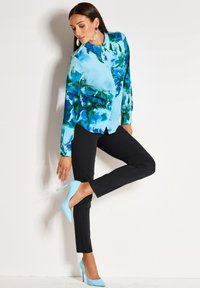 Woman wearing a blue floral blouse, black pants, and light blue high heels, lifting one foot while touching her shoe against a white wall.