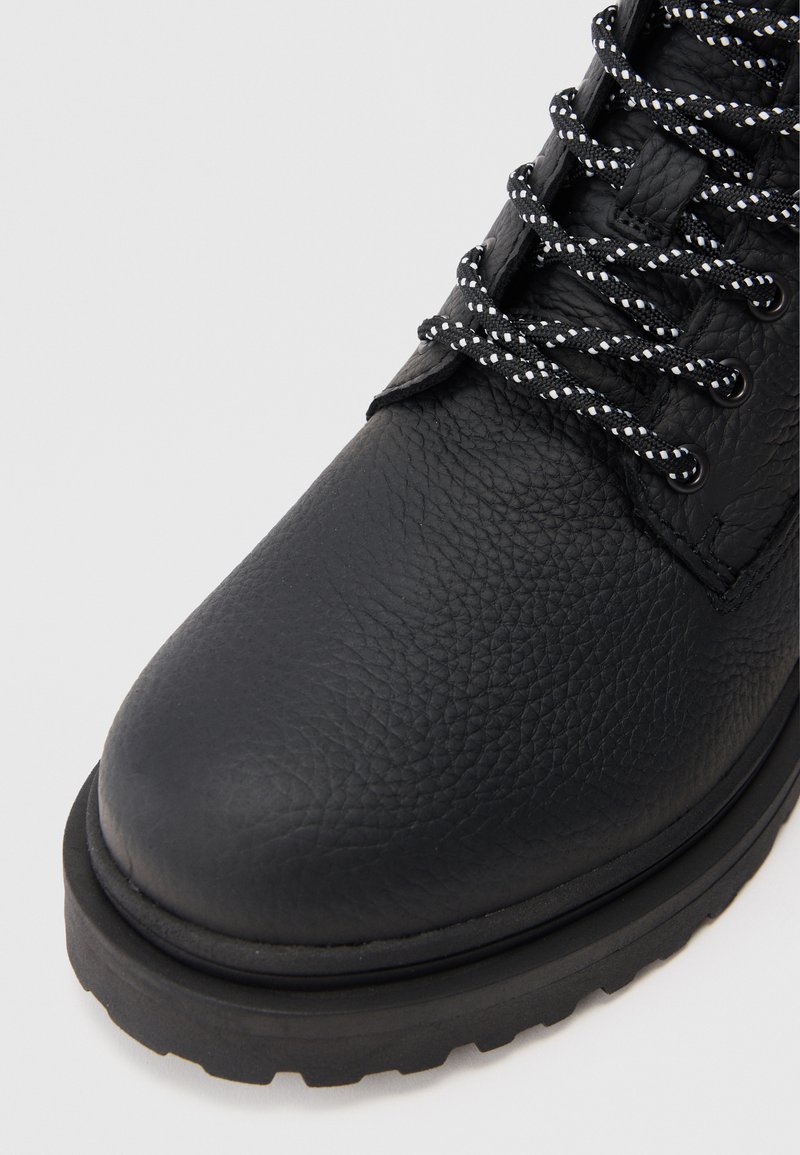 Black leather ankle boot with textured finish, round toe, black and white patterned laces, and sturdy black rubber outsole.