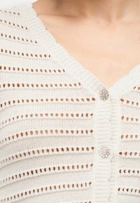 Κοντινή λήψη λευκού πλεκτού ζακέτας με οριζόντιες ρίγες με οπές και δύο στρογγυλά κουμπιά με στρας στο στήθος.