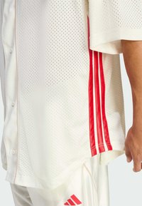 T-shirt atletico bianco perforato con strisce rosse sui lati, chiusura con bottoni sul davanti e maniche corte. Il tessuto sembra traspirante e leggero.