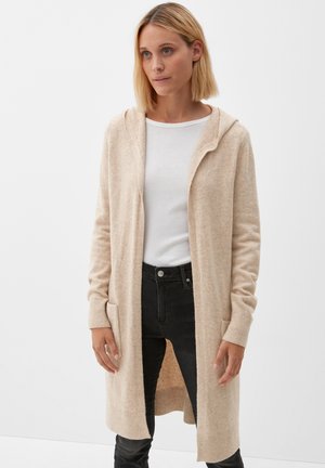 Lichtbeige cardigan met een capuchon, gebreide textuur, voorzakken, open voorkant en geribde manchetten, gecombineerd met een witte top en zwarte jeans.