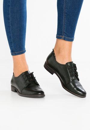 Zapatos de cuero negro con un acabado suave, detalle de cordones en la parte delantera y un tacón bajo. Llevados con jeans ajustados de color azul oscuro.