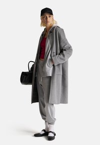 Cappotto di lana grigio sopra un felpa grigia con zip e pantaloni della tuta abbinati. Cappello nero, piccola borsa nera e scarpe nere con calzini bianchi.