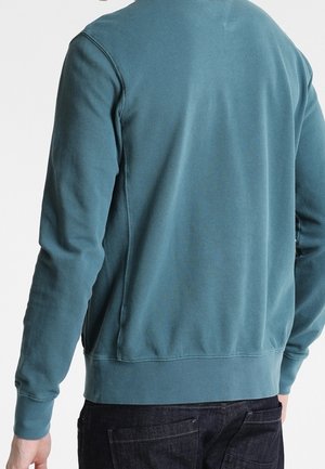 Mann trägt blaues Sweatshirt mit langen Ärmeln und dunkle Jeans, von hinten vor schlichtem weißen Hintergrund.