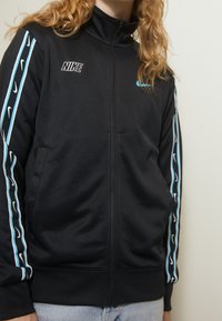 Nike svart zip-up jacka med Aqua-ränder längs ärmarna och Nike-loggan i vitt på bröstet, tillverkad av mjukt, stretchigt material.