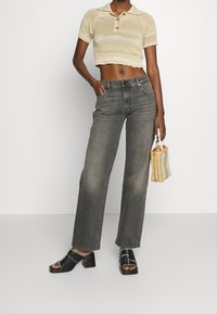 Jeans grigi alti in vita con una vestibilità comoda, abbinati a un top corto beige chiaro dalla trama particolare e sandali neri con tacco a blocco, mentre tiene in mano una borsa a quadri.