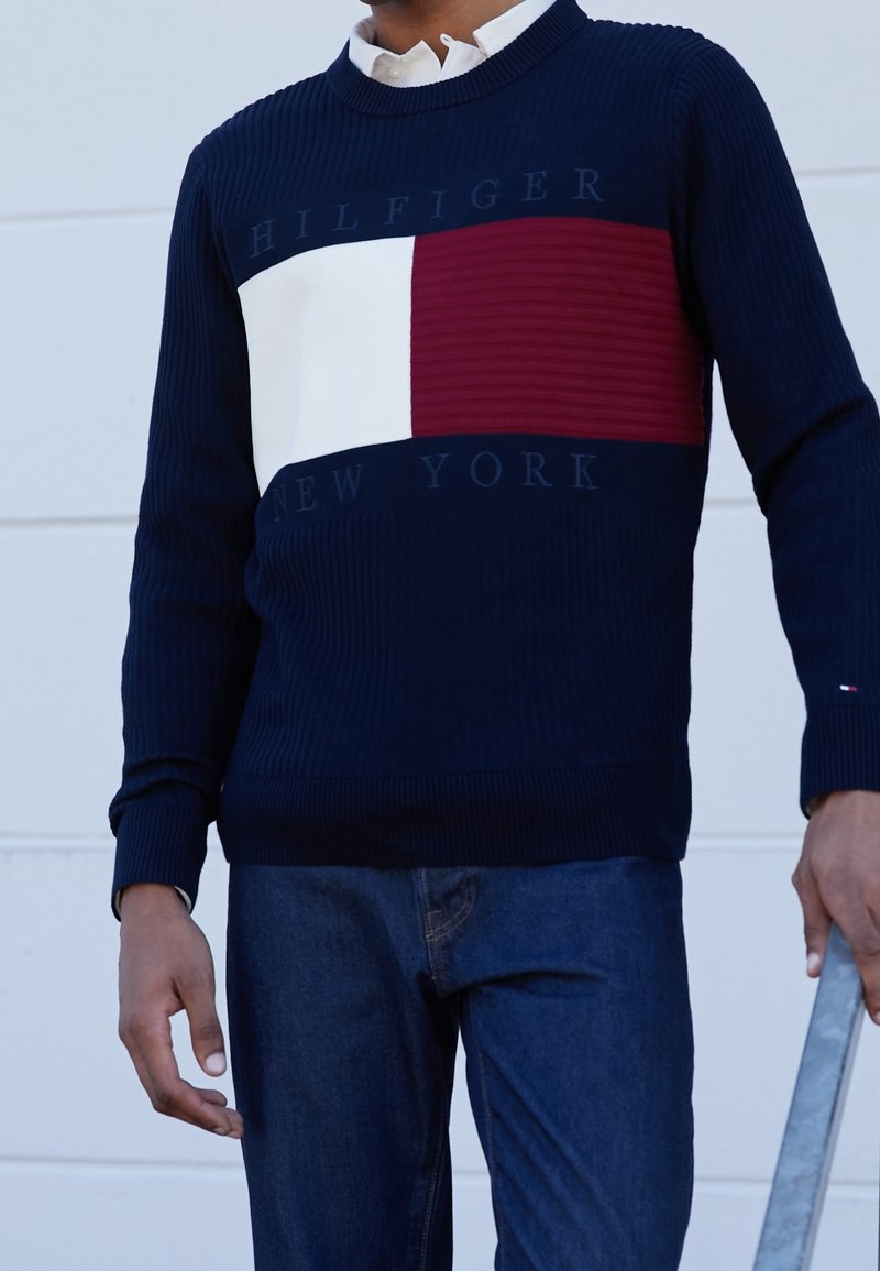 Pull en maille côtelée bleu marine avec un grand panneau rectangulaire blanc et rouge. Logo "HILFIGER NEW YORK" affiché sur la poitrine.