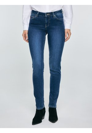 Moteris dėvi aptemptus tamsiai mėlynus džinsus, baltą ilgomis rankovėmis marškinius ir juodus kulkšninius batus, stovinti priešais paprastą foną.