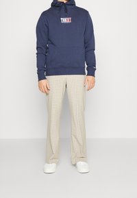 Navy hooded sweatshirt with a central logo and front pocket, paired with light brown checked trousers. White sneakers complete the outfit.