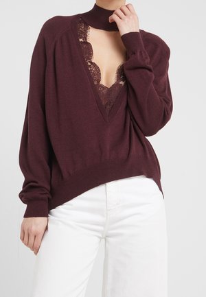 Frau trägt burgunderfarbenen V-Ausschnitt-Pullover mit Spitzendetails und hohem Kragen, kombiniert mit hoch taillierter weißer Hose vor einfachem Hintergrund.