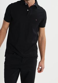 Musta polo-paita lyhyillä hihoilla, kauluksella ja kahdella napilla. Siinä on pieni logo rinnassa ja sileä puuvillapinta.