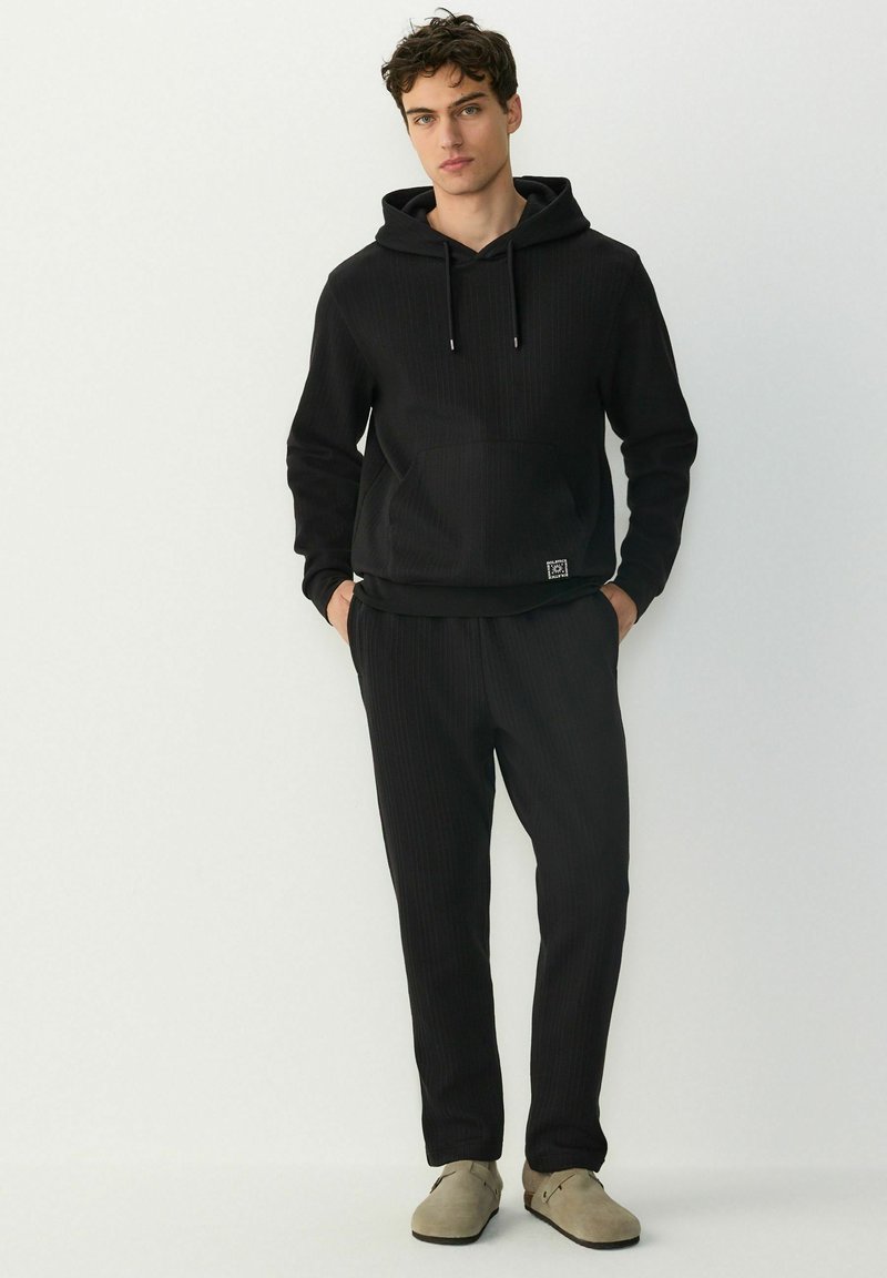 Sudadera negra con textura acanalada, con un bolsillo frontal y cordones, combinada con pantalones a juego también acanalados. Tejido suave con un corte relajado, llevada con zapatos ligeros.