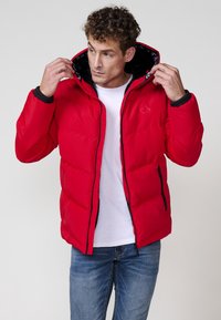 Chaqueta acolchada roja con capucha, que cuenta con bolsillos frontales con cremallera y un forro negro. La chaqueta tiene un diseño acolchado con una forma ajustada.
