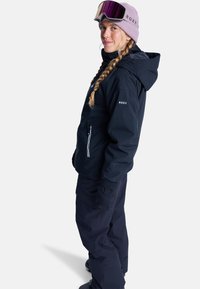 Woman wearing black ski jacket and pants, pink beanie with ski goggles, standing sideways with hands in pockets on white background.