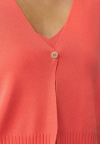 Korallfarbener Cardigan mit V-Ausschnitt, der über einen einzelnen Knopfverschluss und einen gerippten unteren Saum verfügt. Hergestellt aus einem weichen, glatten Strickmaterial.