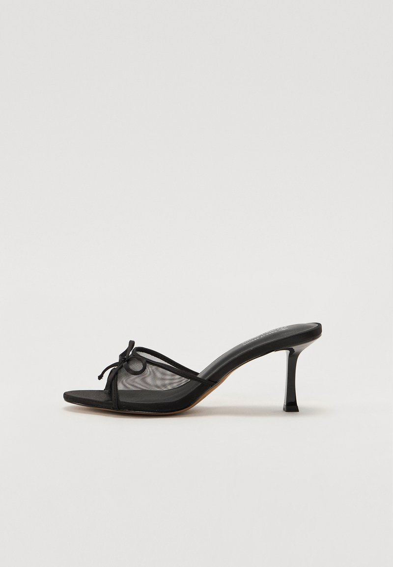 Zwarte heeled mule met een bovenwerk van gaas, dunne bandjes en een decoratieve knoop. Heeft een slanke, puntige hak en een gestructureerde binnenzool.
