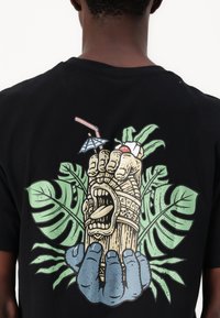 Zwarte t-shirt met een kleurrafische afbeelding van een tiki-mok vastgehouden door een blauwe hand, omringd door groene bladeren en een drinkparaplu.