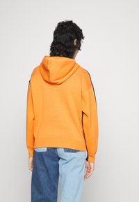 Person with dark curly hair wearing an orange hooded sweatshirt and two-tone blue jeans facing away against a plain light background.