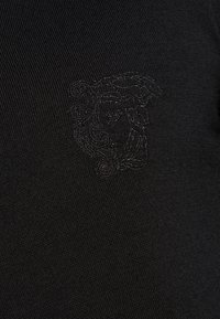 Czarna tkanina teksturowana z subtelnym haftowanym wzorem przedstawiającym twarz na froncie. Tkanina ma delikatny, ribbowany wzór.