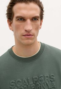 Groene sweatshirt met een getextureerd logo aan de voorkant. Heeft een geribbelde kraag en zachte stof. Het ontwerp is minimalistisch en modern.