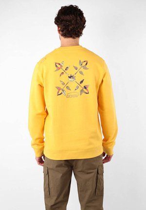 Sweatshirt jaune avec un motif floral graphique au dos, comprenant diverses formes et couleurs. Poignets et ourlet côtelés. Matériau en coton.