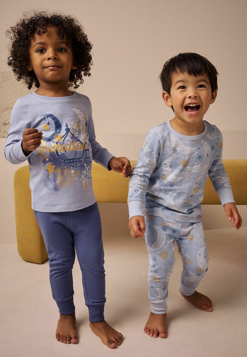 Due bambini scalzi che indossano pigiami blu a tema Ramadan stanno in piedi e sorridono all'interno vicino a una panchina color senape.