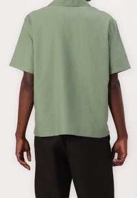 Camisa de manga corta en tejido de color verde claro texturizado, con cuello puntiagudo y dobladillo recto, acompañada de pantalones negros.
