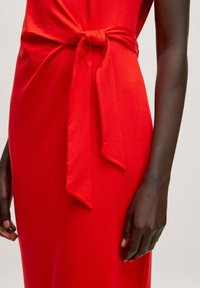 Rotes Kleid mit einem gebundenen KnotenDetail an der Taille, aus glattem Stoff mit fließendem Design, das klare Linien und eine figurbetonte Silhouette zeigt.