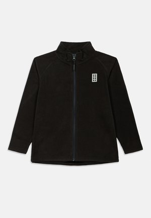 Črna flis jakna z visokim ovratnikom, polnim zadrgo in raglan rokavi. Ima majhen bel logotip na levi prsni strani. Mehka tekstura.