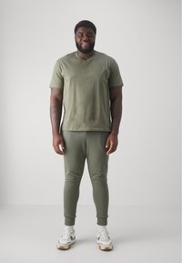 Mann steht, trägt ein olivgrünes kurzärmliges Shirt und Jogginghosen, weiße Socken und weiße Sneakers mit grünen Akzenten, vor einem schlichten hellen Hintergrund.