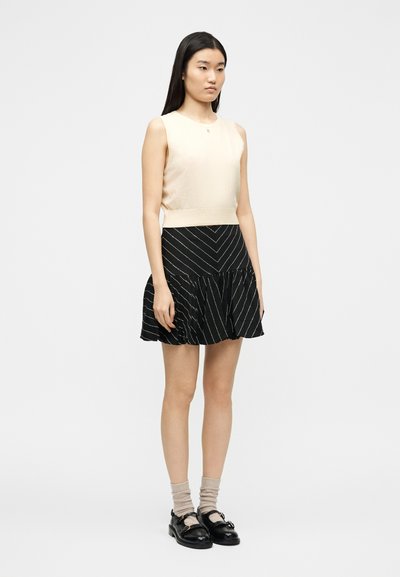 Γυναίκα με μακριά σκούρα μαλλιά που φοράει μπεζ αμάνικο τοπ, φούστα με ασπρόμαυρες ρίγες,  μπεζ κάλτσες και μαύρα μοκασίνια, στεκόμενη σε ουδέτερη στάση.