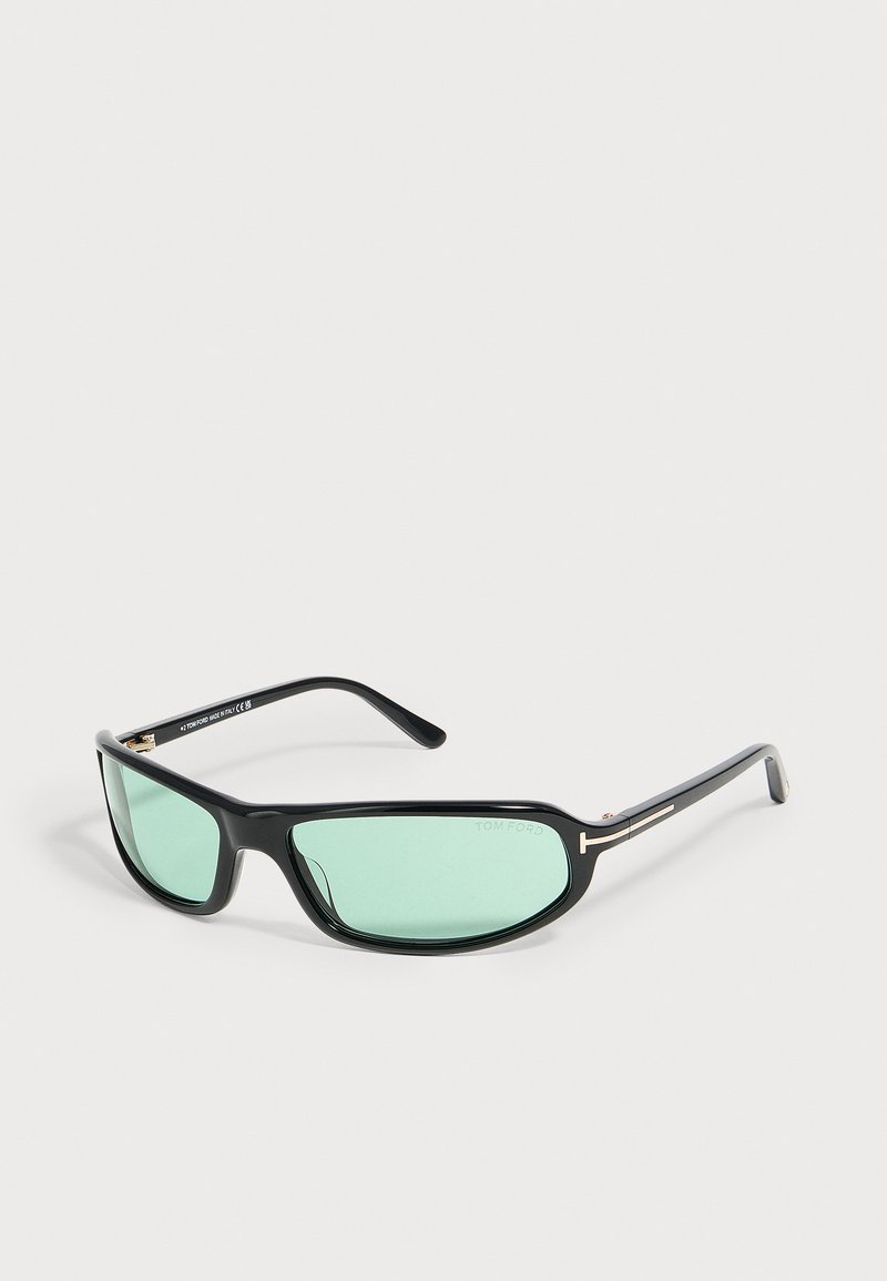 Lunettes de soleil à monture noire avec des verres vert clair et des détails métalliques sur les branches, posées sur un fond neutre uni.