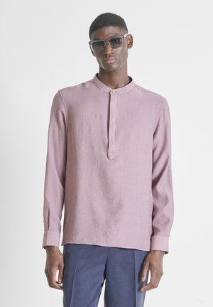 Jonge man draagt een oversized paarse overhemd met mandarijnkraag, marineblauwe broek en donkere zonnebril tegen een effen witte achtergrond.