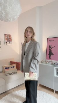 Sacou gri în carouri, croială oversized, purtat peste un top alb. Pantaloni negri cu picior larg, ținând o poșetă mică bej. Decor minimalist în fundal.