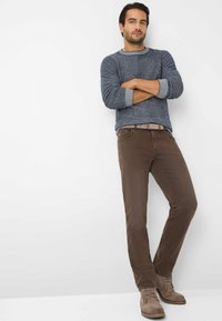 Uomo appoggiato a un muro con le braccia incrociate, che indossa un maglione grigio, pantaloni marroni, una cintura beige e stivali beige con lacci.