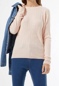 Lichtroze kabelgebreide trui met geribde zoom en manchetten, gedragen met een spijkerjack, tegen een neutrale achtergrond.